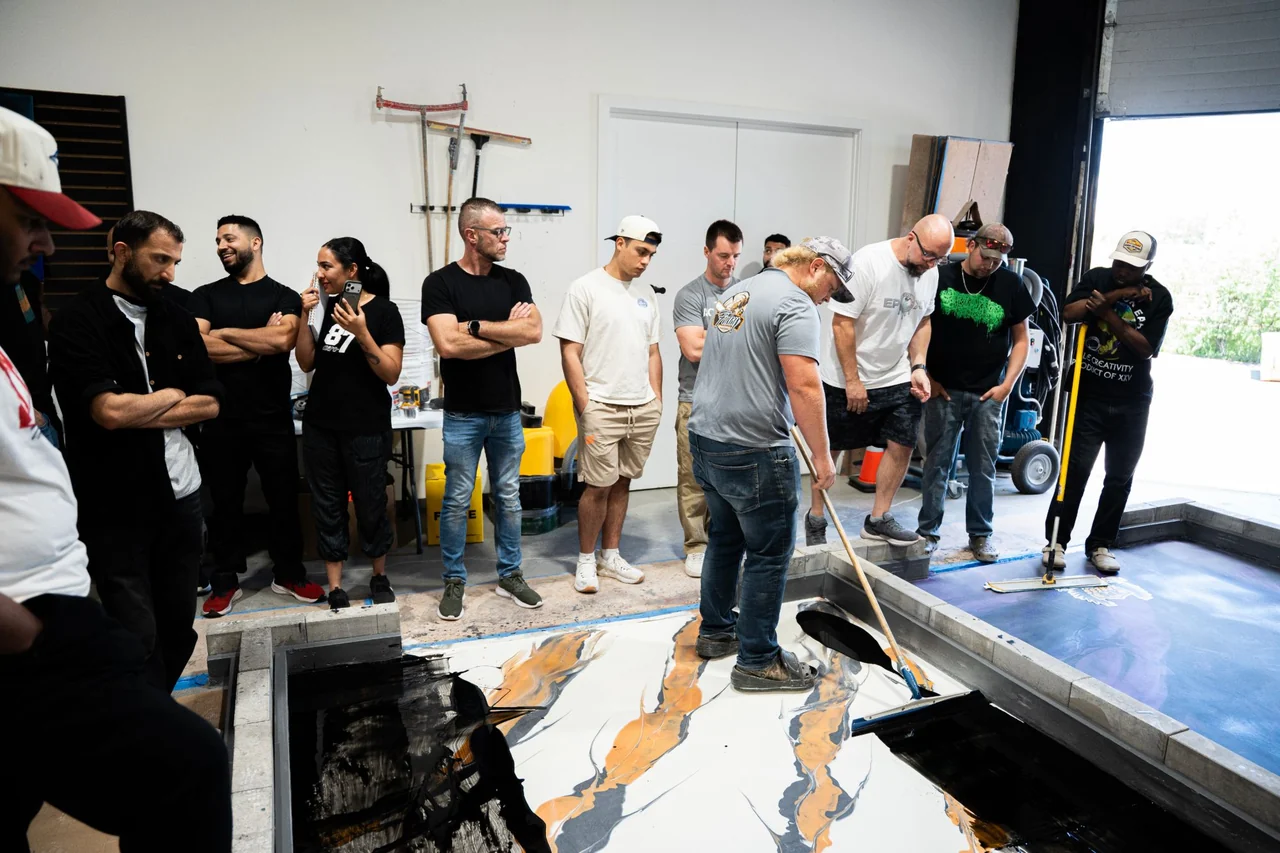 A student spreads a metallic epoxy pour with a squeegee as the instructor and class watch